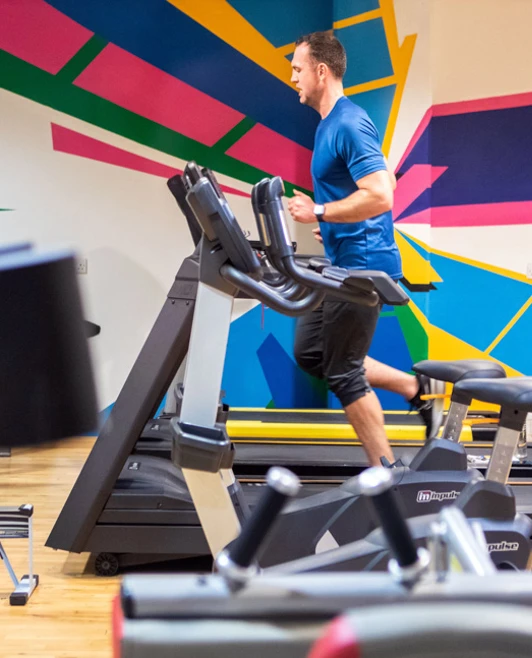 A man running on a treadmill in a brightly coloured gym with various fitness machines. A man running on a treadmill in a brightly coloured gym with various fitness machines.